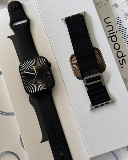 A black smartwatch with a sunburst-patterned black display and a black sport band, shown alongside a black woven loop watch band on a white box. The brand name 'unibonds.