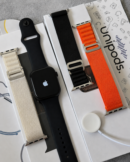 Apple Watch with a black band laid on top of white UniPods charging/accessory boxes, alongside two additional woven watch bands in beige and bright orange.