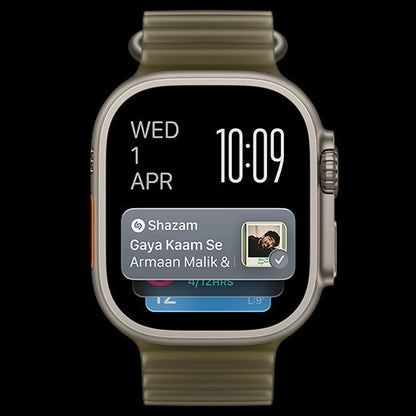 A smartwatch with a metallic silver-toned case and a textured olive green band, shown against a solid black background. The watch face is active with a digital clock showing 10:09 and a notification banner from 'Shazam' displayed on the screen.